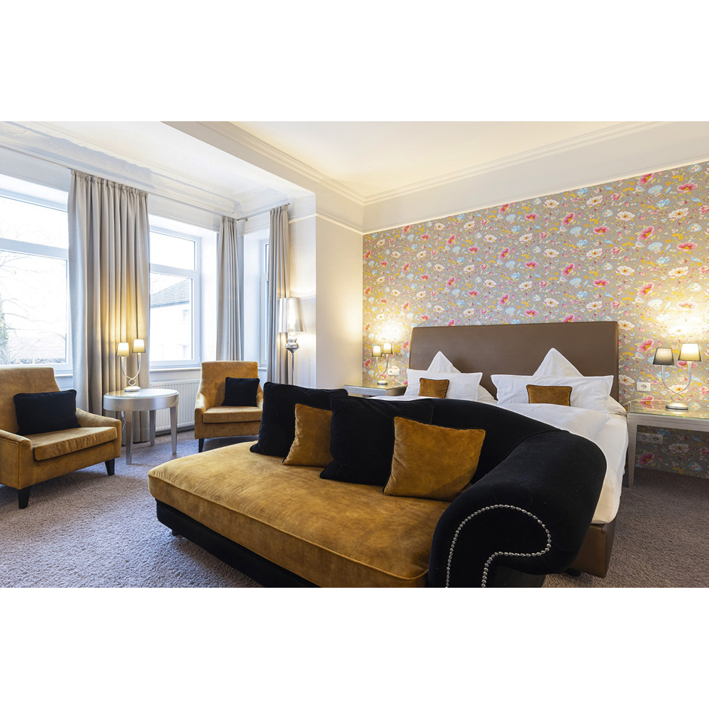 Ein Hotelzimmer mit großem Bett, gelb-schwarzem Sofa, Sesseln, Blumentapete und großen Fenstern mit Vorhängen.