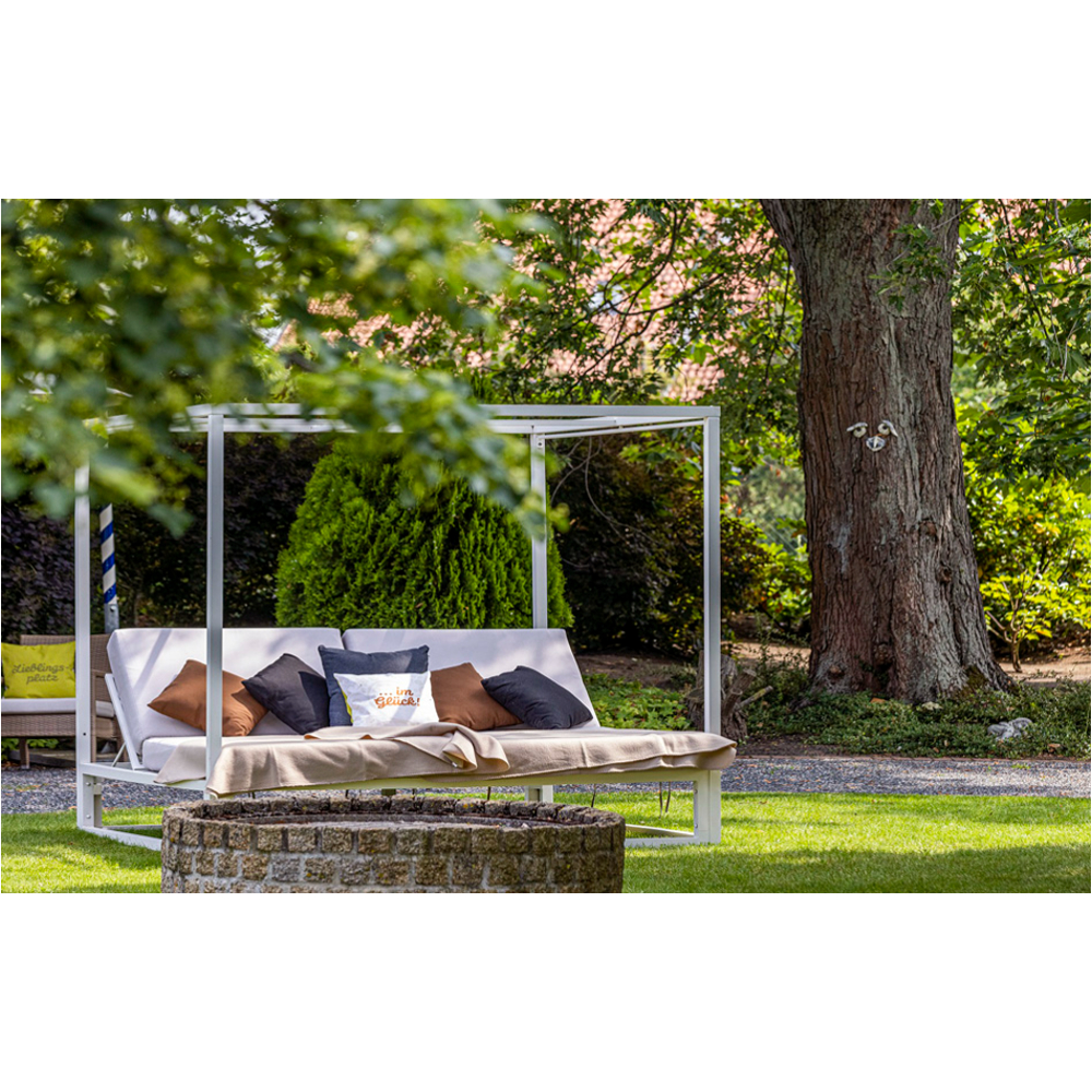 Eine weiß gerahmte Tagesliege im Freien mit Kissen in einem grünen Garten.
