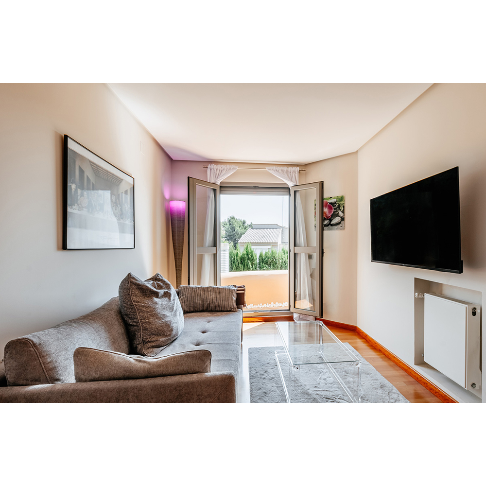Wohnzimmer mit grauem Sofa, Glastisch, Fernseher an der Wand und offenem Balkon mit Blick auf Bäume.