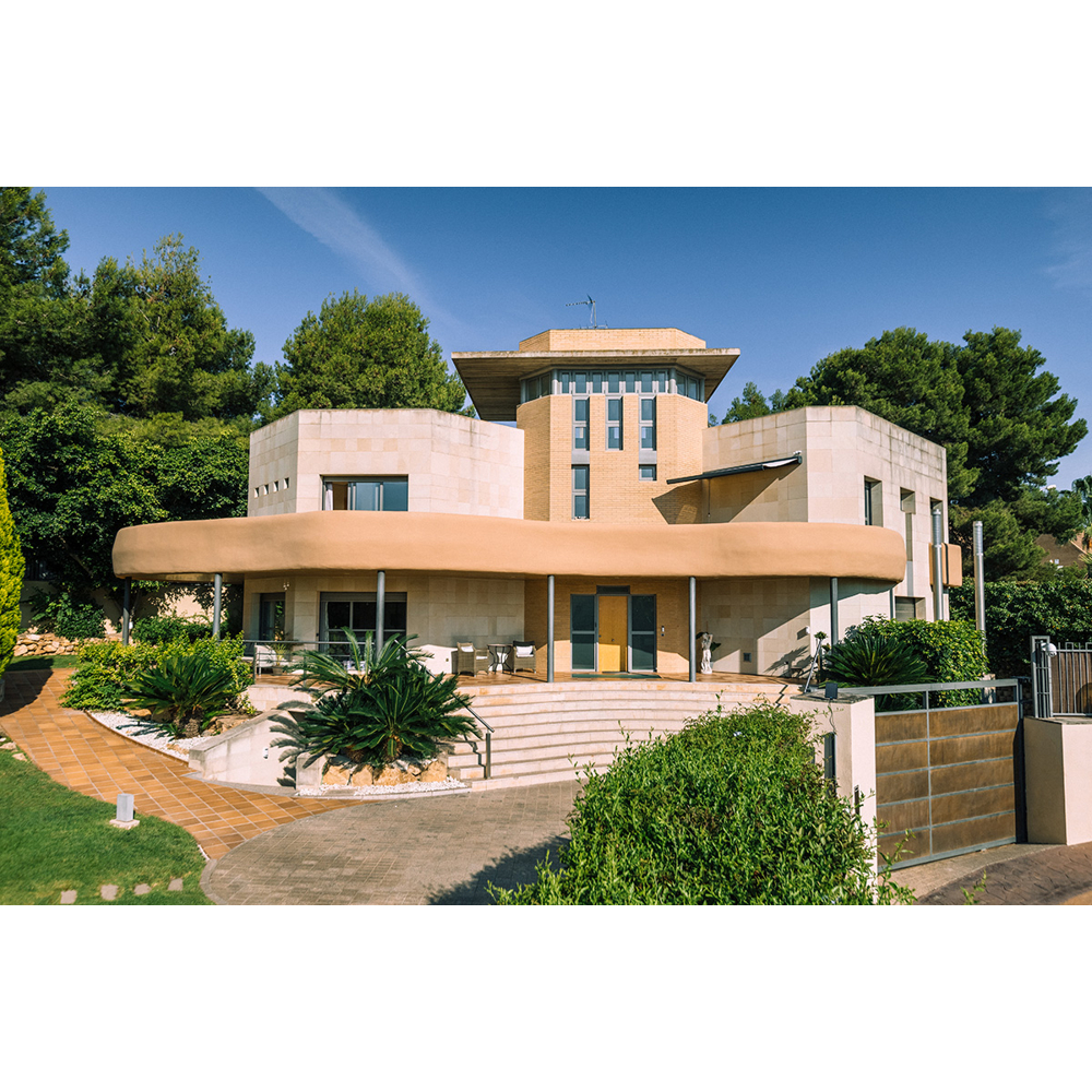 Eine moderne beige Villa mit kreisförmiger Terrasse, umgeben vongrünem Garten und Bäumen unter blauem Himmel.