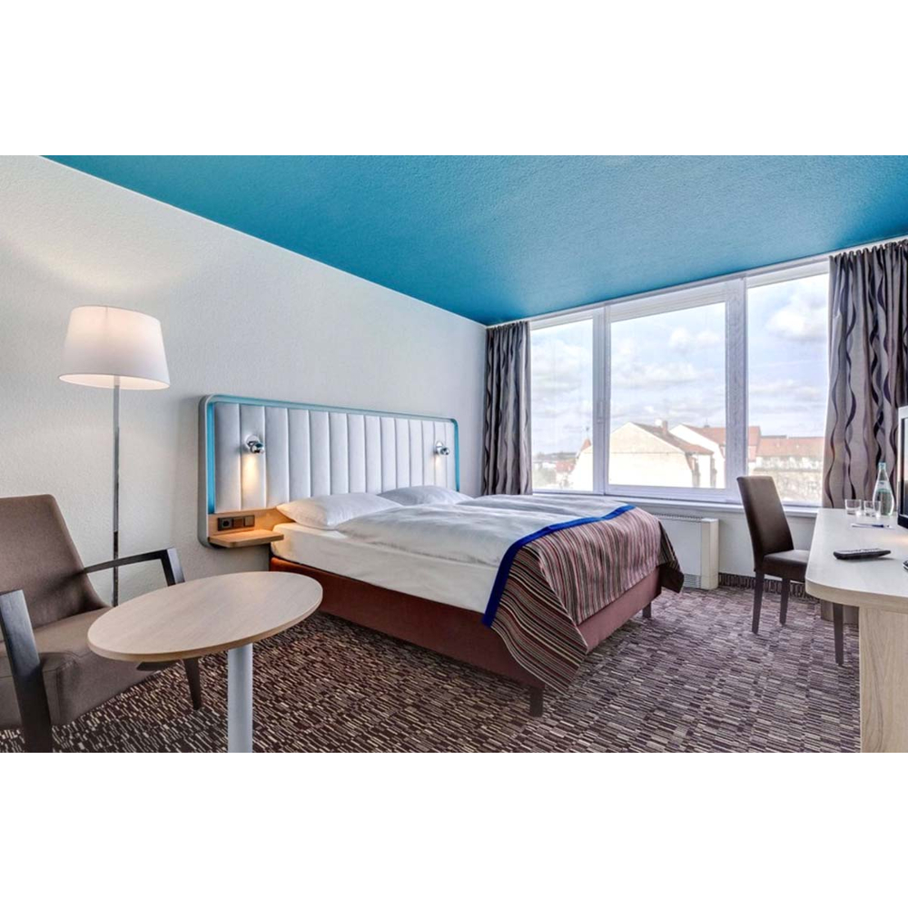 Ein helles Hotelzimmer mit weißem Bett, blauer Decke, Stehlampe, Sesseln und großen Fenstern mit Stadtblick.