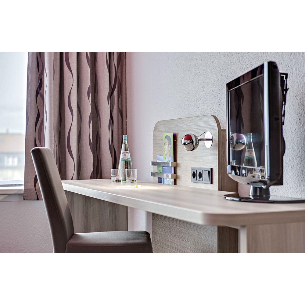 Ein Schreibtisch mit Stuhl, Fernseher, Flasche und Gläsern steht vor einem Fenster mit gemustertem Vorhang.