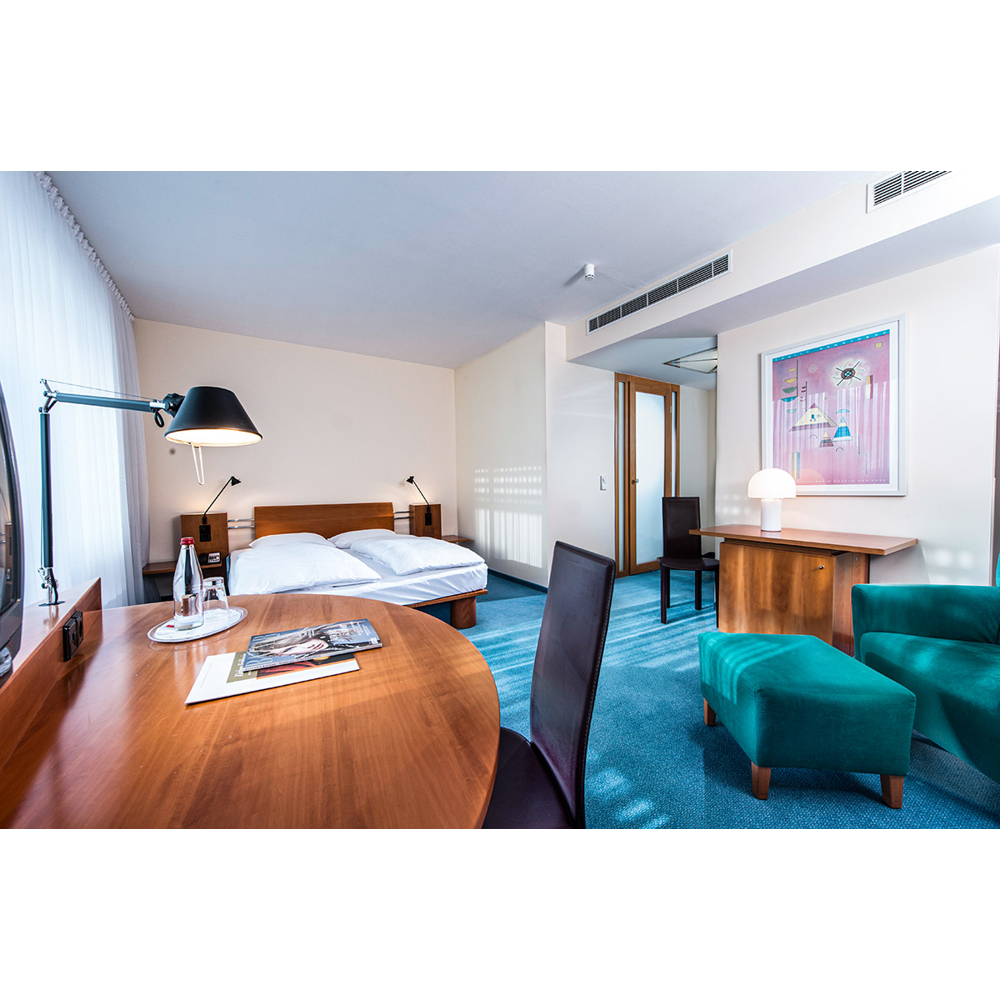 Ein modernes Hotelzimmer mit Holzbett, blauem Teppich, Schreibtisch und einer grünen Sitzecke mit Hocker.