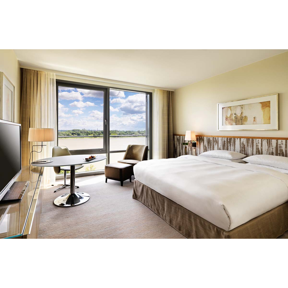 Moderne Hotelzimmer mit großem Bett, Schreibtisch, Sessel und Fensterblick auf einen Fluss unter bewölktem Himmel.