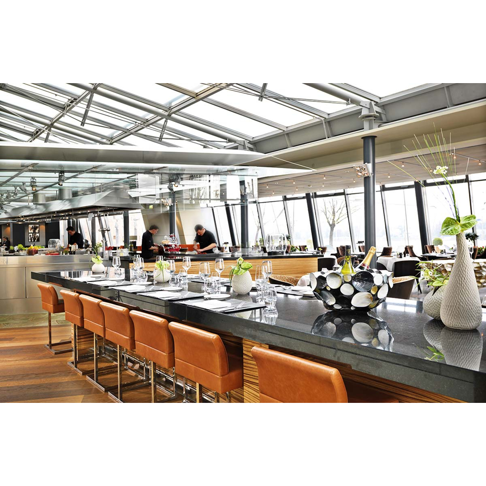 Ein modernes Restaurant mit langer schwarzer Theke, braunen Barhockern, Glasdach und Köchen im Hintergrund.