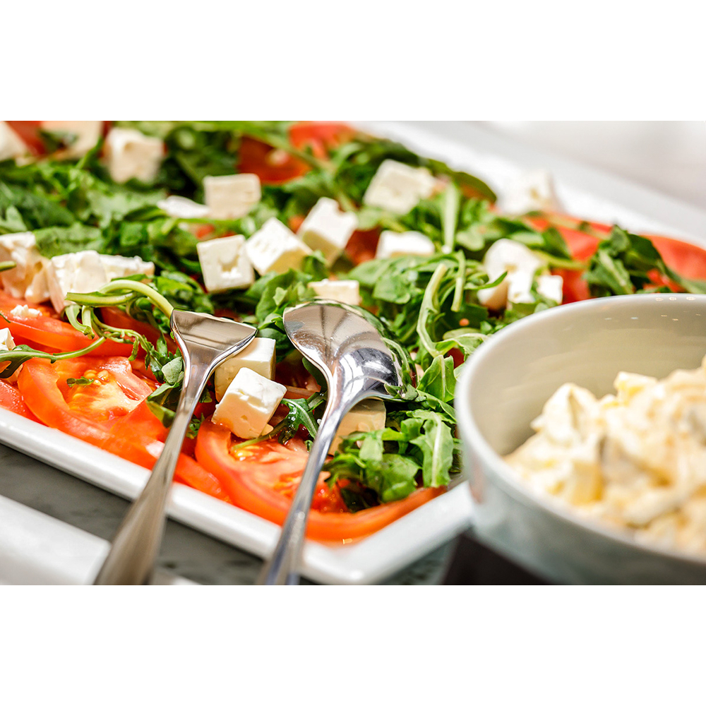 Ein frischer Salat mit Tomaten, Rucola und Feta-Würfeln auf einer weißen Platte mit Vorlegebesteck.