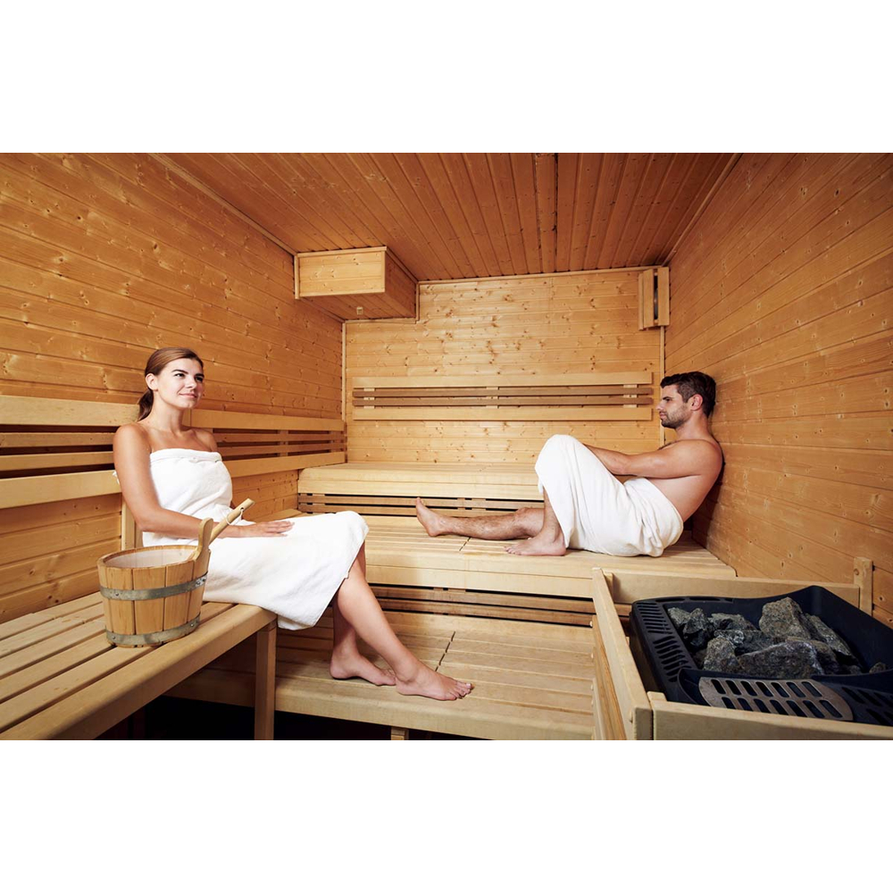Ein Mann und eine Frau sitzen in Handtüchern in einer holzverkleideten Sauna, neben einem Eimer und Ofen.