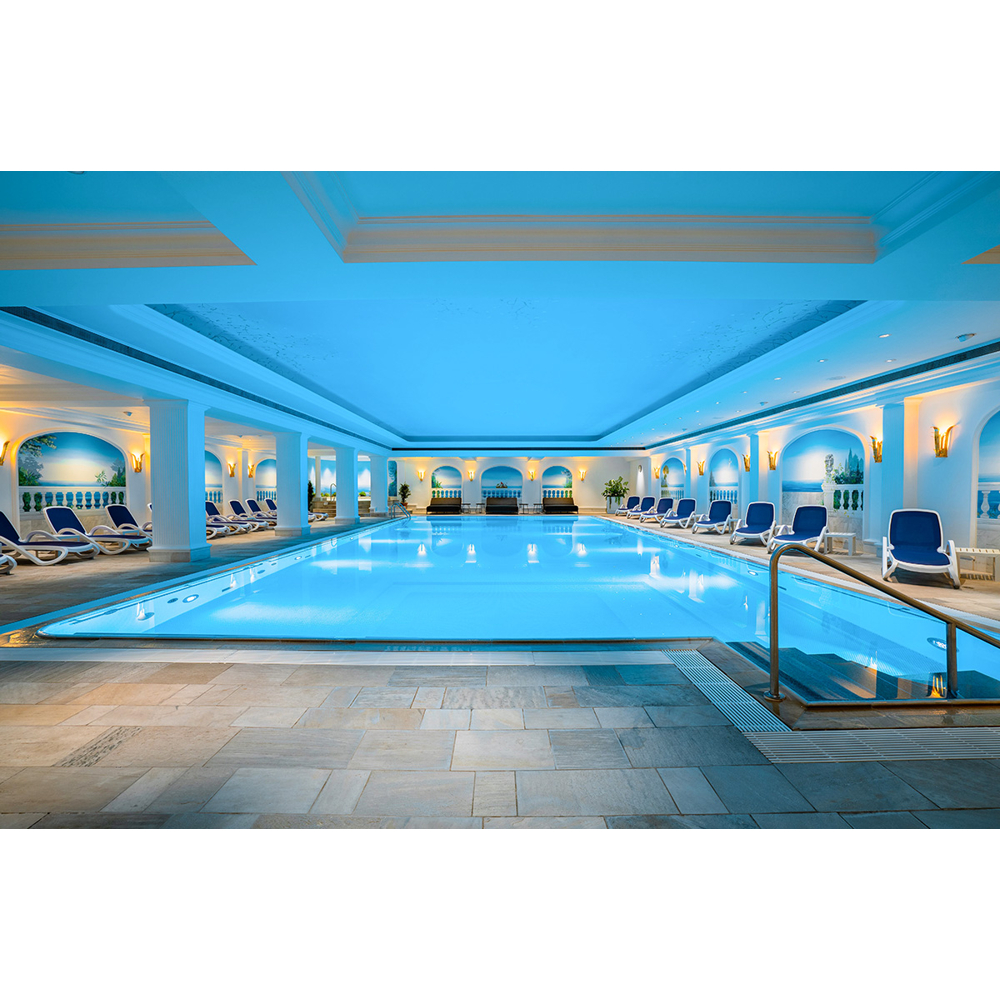 Ein großes, blaues Hallenbad mit Säulen, blauer Beleuchtung und Liegestühlen an den Seiten.