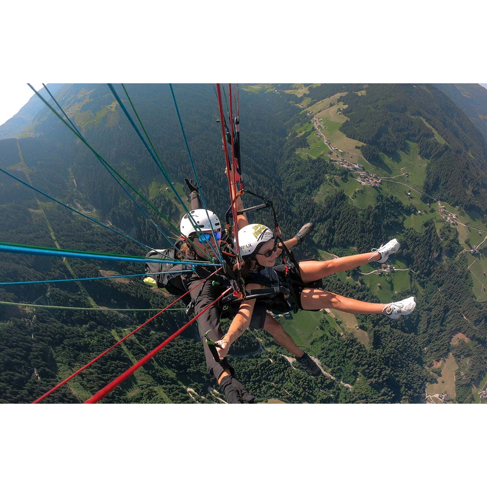 Zwei Personen paragliden über ein bewaldetes Tal mit bunten Seilen im Vordergrund.