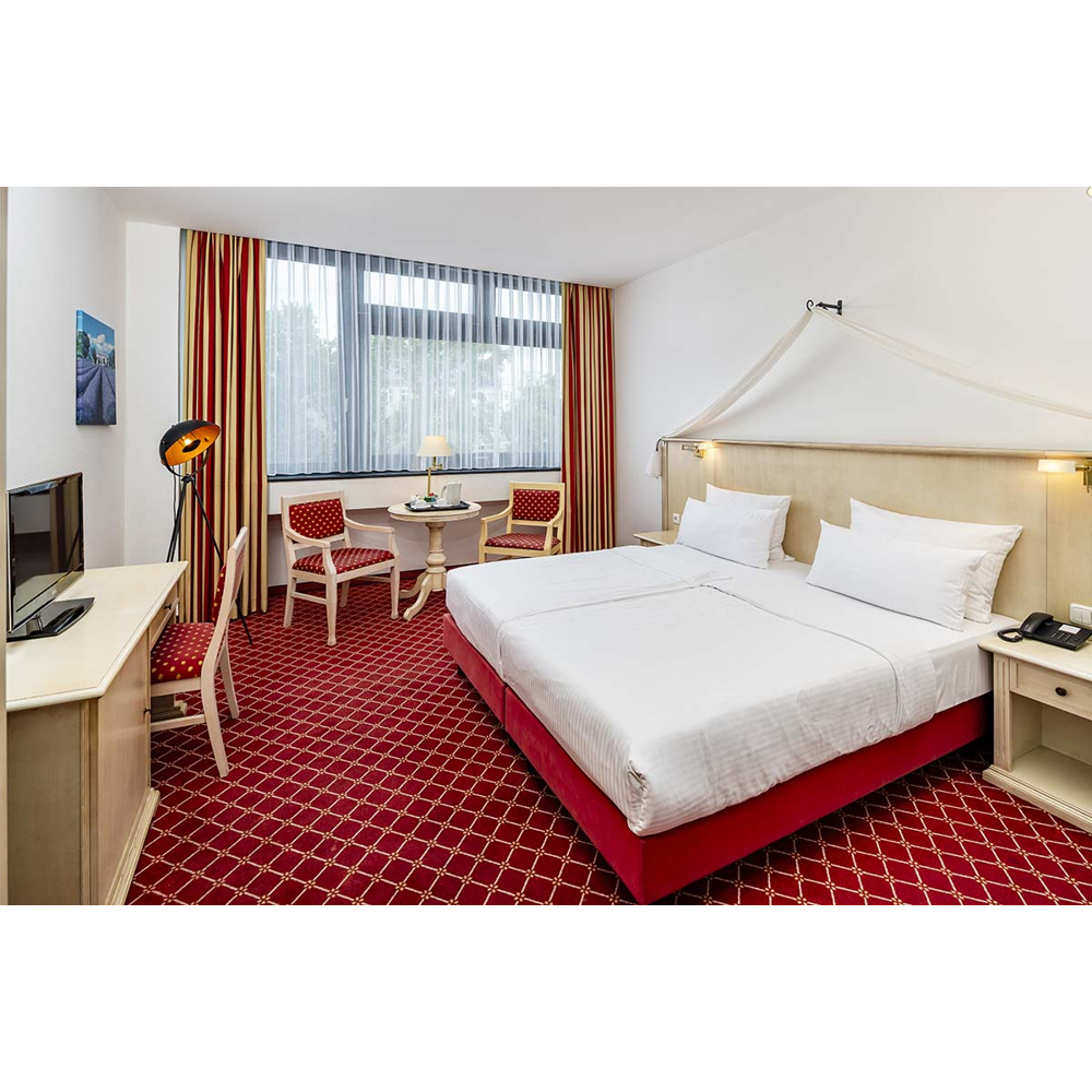 Ein Hotelzimmer mit rotem Teppich, großem Doppelbett mit weißer Bettwäsche, Fenstern mit roten und goldenen Vorhängen und einer Sitzecke.