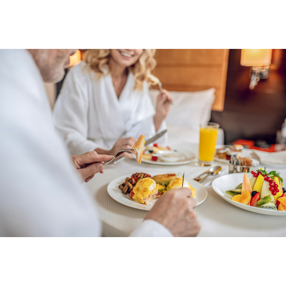 Ein Paar in weißen Bademänteln genießt ein Frühstück im Bett mit Eiern und frischem Obst.