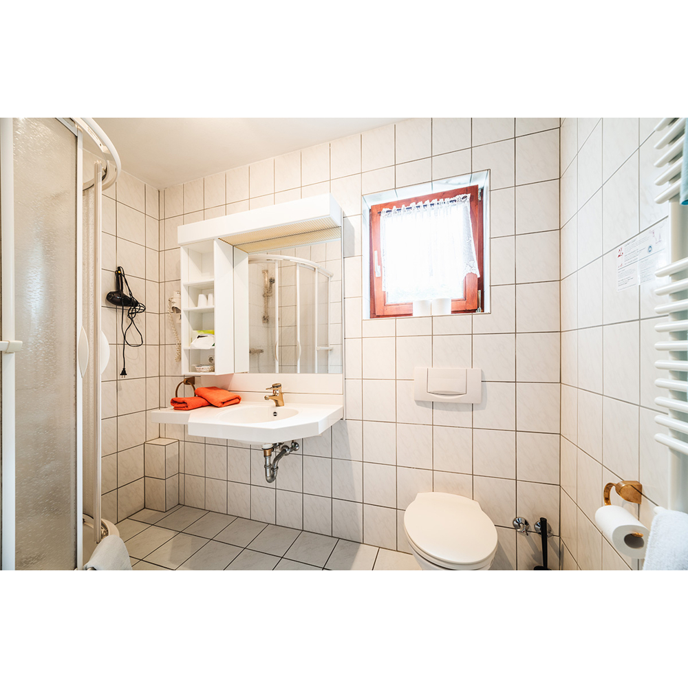 Ein helles Bad mit weißen Fliesen, Waschbecken, orangefarbenem Handtuch, Toilette, Duschkabine und kleinem Fenster.