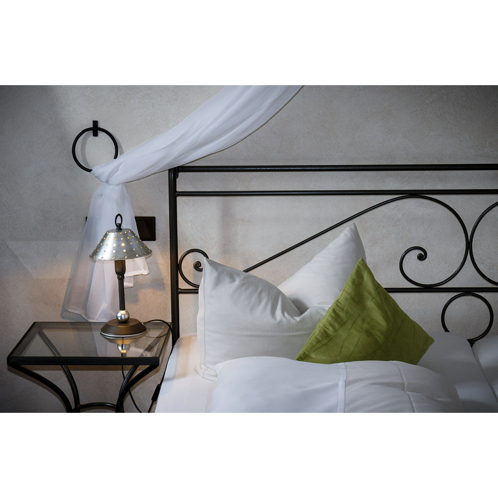 Ein Bett mit dunklem Metallrahmen, weißen Kissen, einem grünen Kissen und einer Nachttischlampe daneben.