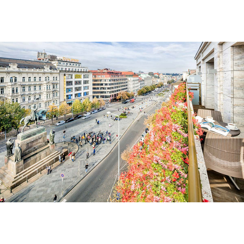Erhöhte Stadtsicht mit geschäftigem Platz, großer Reiterstatue auf Sockel und Balkon voller roter Blumen.