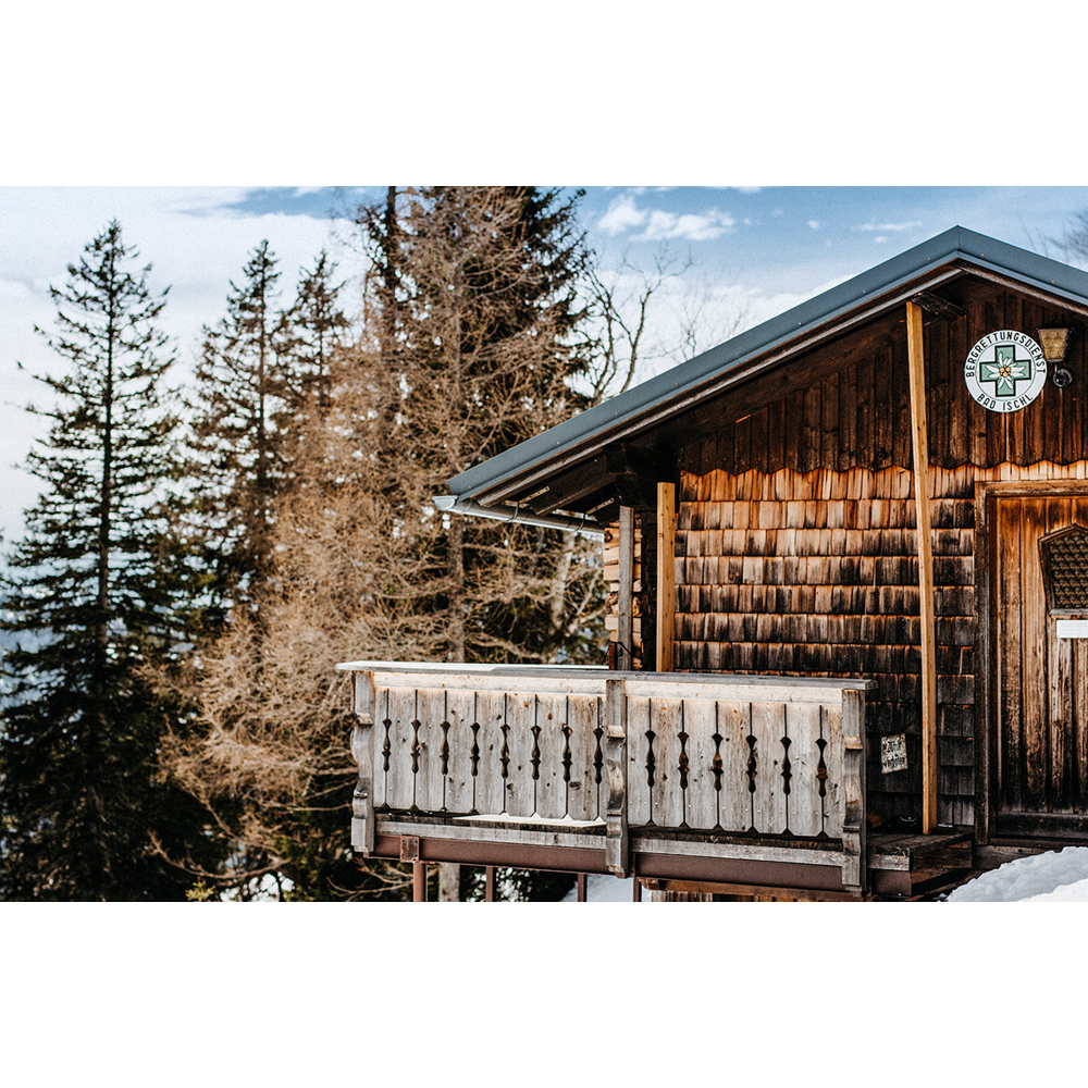 Eine Holzhütte mit Balkon steht in einer winterlichen Berglandschaft mit vielen Tannen.
