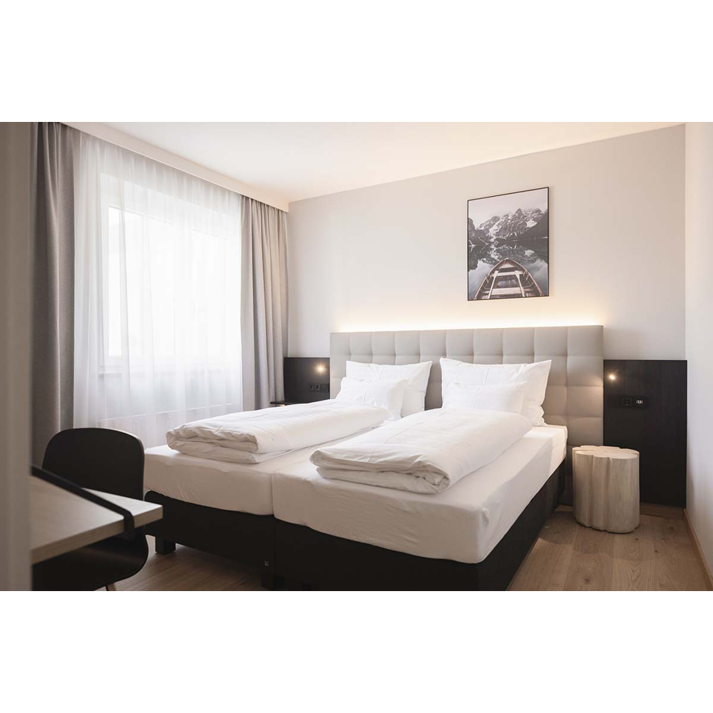 Ein modernes Hotelzimmer mit Doppelbett, weißer Bettwäsche, großem Fenster mit Vorhängen und einem Naturbild an der Wand.