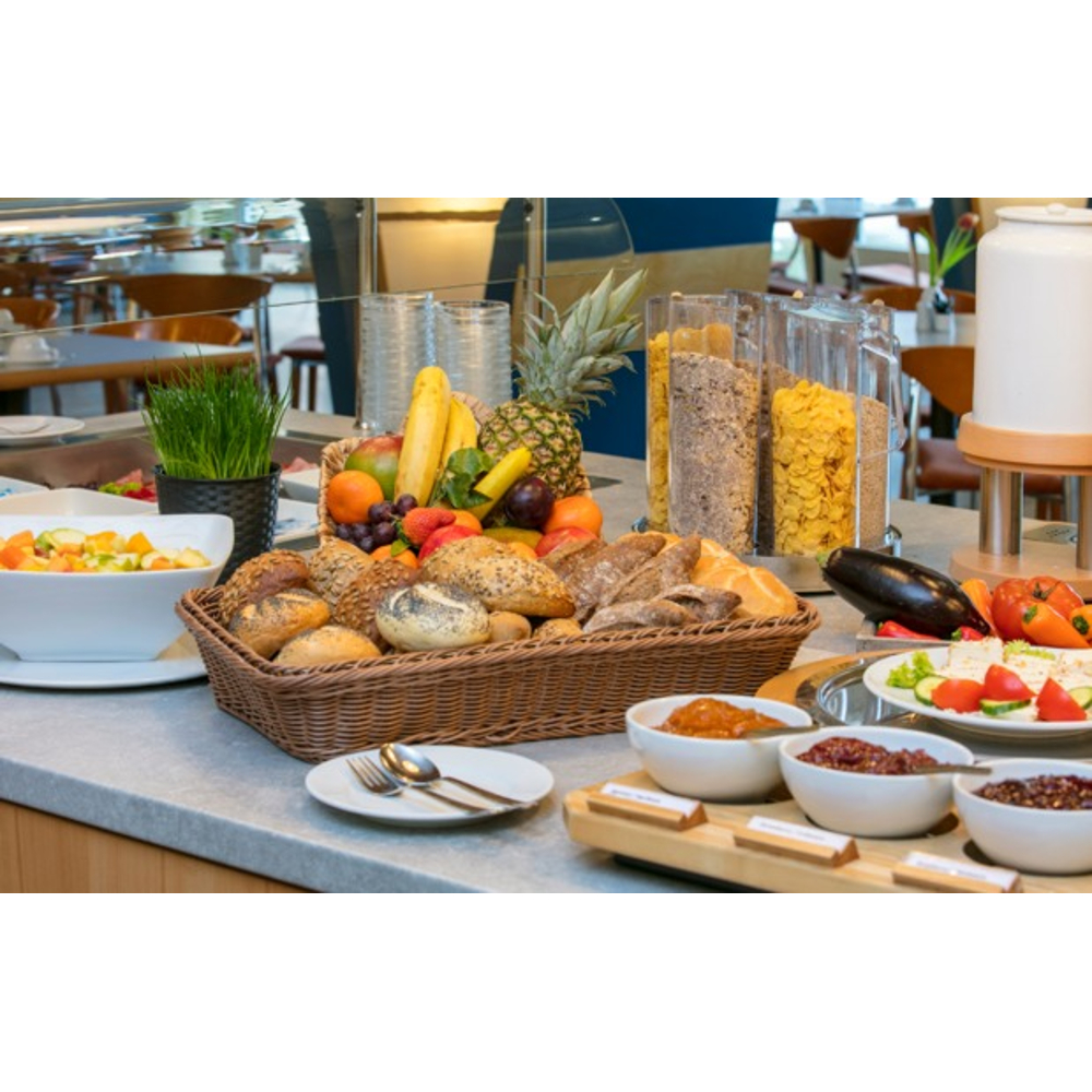 Ein Frühstücksbuffet mit frischem Obst, Brot in einem Korb, Müslispendern und verschiedenen Aufstrichen.