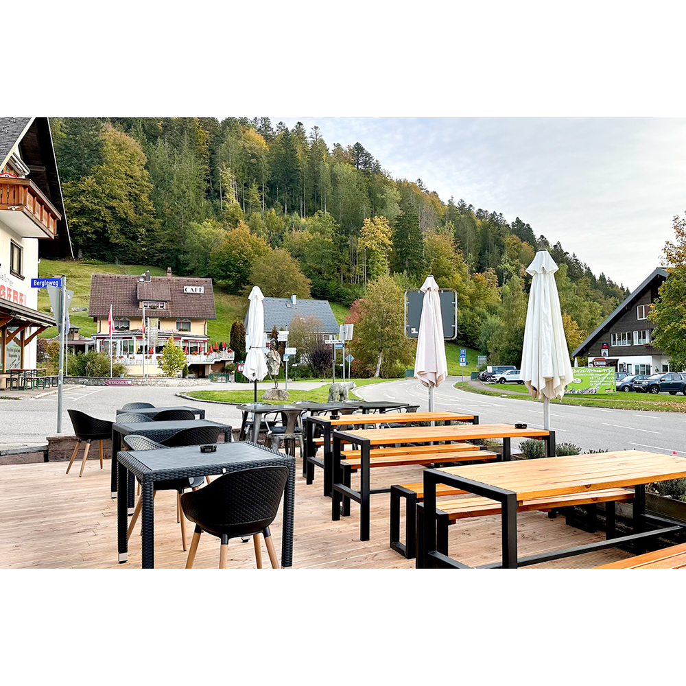 Café-Terrasse im Freien mit Holztischen, schwarzen Stühlen und weißen Sonnenschirmen mit Blick auf einen waldigen Berg.