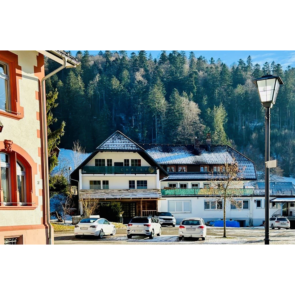 Vier parkende Autos vor einem schneebedeckten Chalet mit Hintergrund eines grünen Nadelwaldbergs unter blauem Himmel.