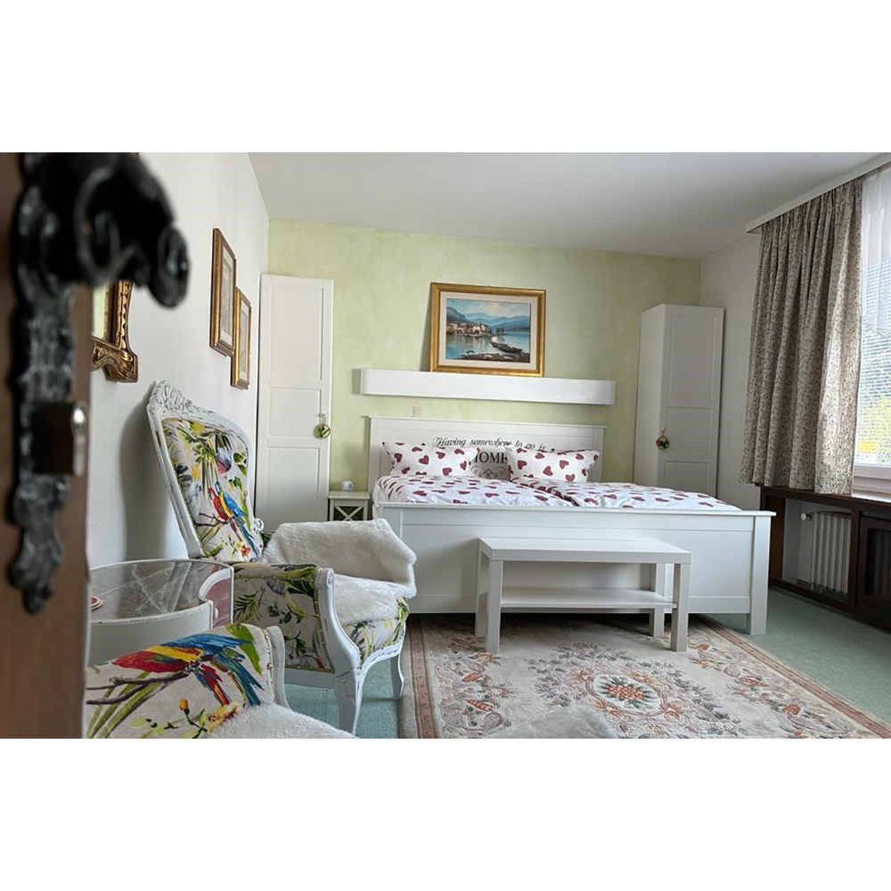 Ein gemütliches Schlafzimmer mit weißem Bett, gemustertem Sessel und gemustertem Teppich auf grünem Boden.