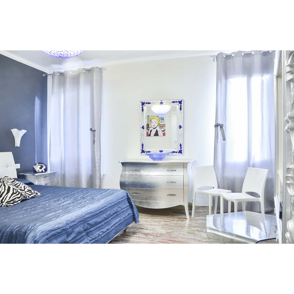 Schlafzimmer mit blauem Bett, silberner Kommode, Kunstspiegel, zwei weißen Stühlen und grauen Vorhängen.