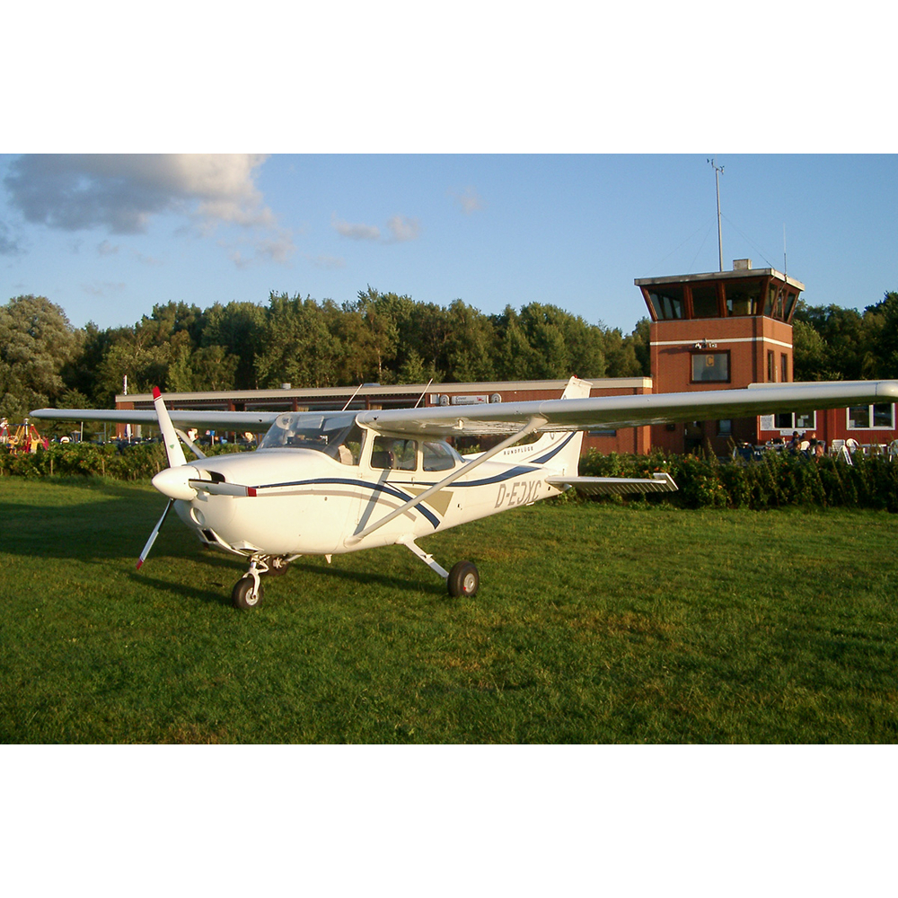 Ein kleines weißes Propellerflugzeug parkt auf einer grünen Wiese vor einem Backsteingebäude.