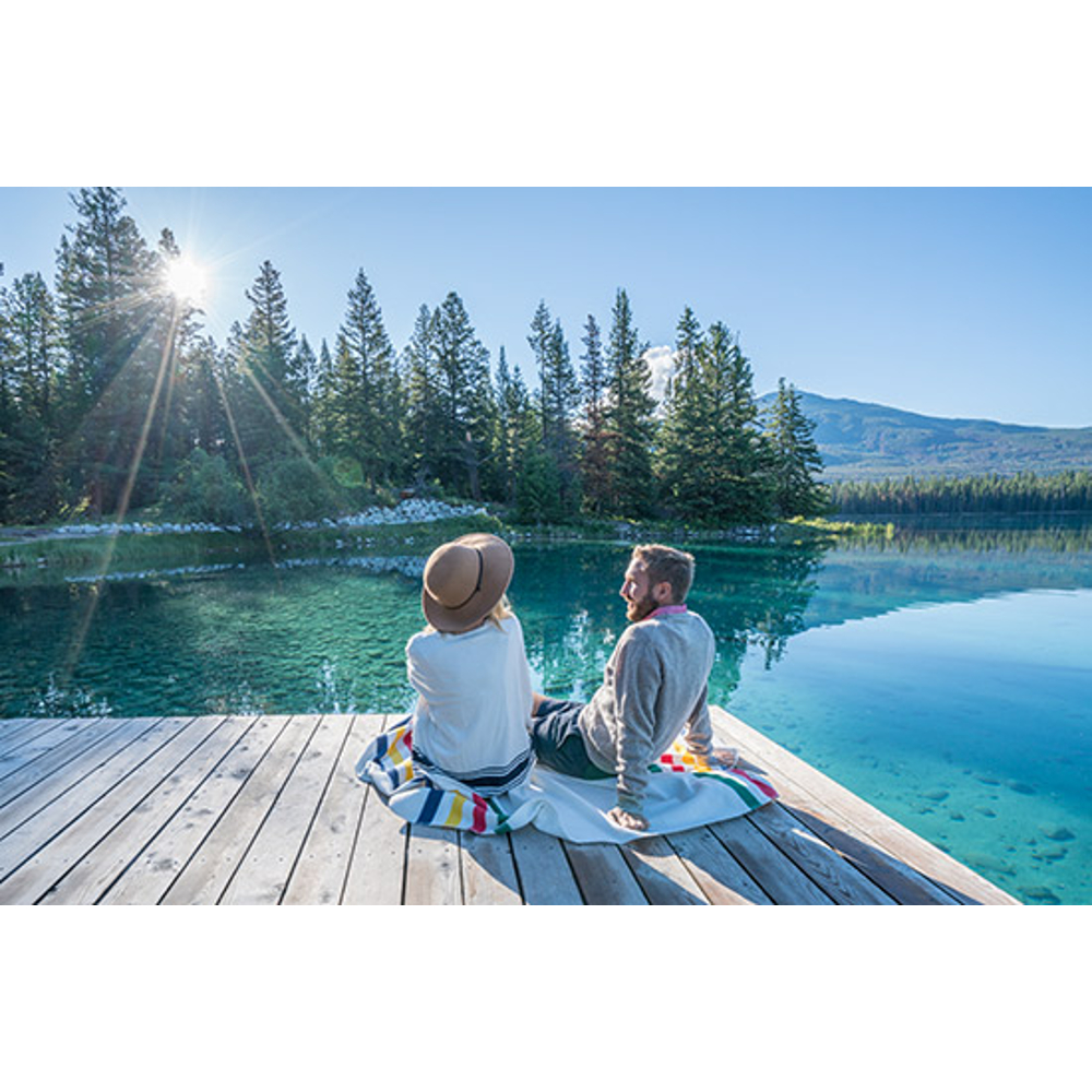 Ein Paar sitzt auf einem Holzsteg an einem klaren blauen See, umgeben von Kiefern unter hellem Sonnenschein.