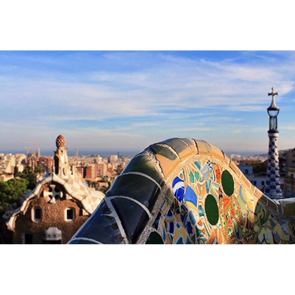Bunter Mosaikbogen im Vordergrund, im Hintergrund der Park Güell und die Skyline von Barcelona unter blauem Himmel.
