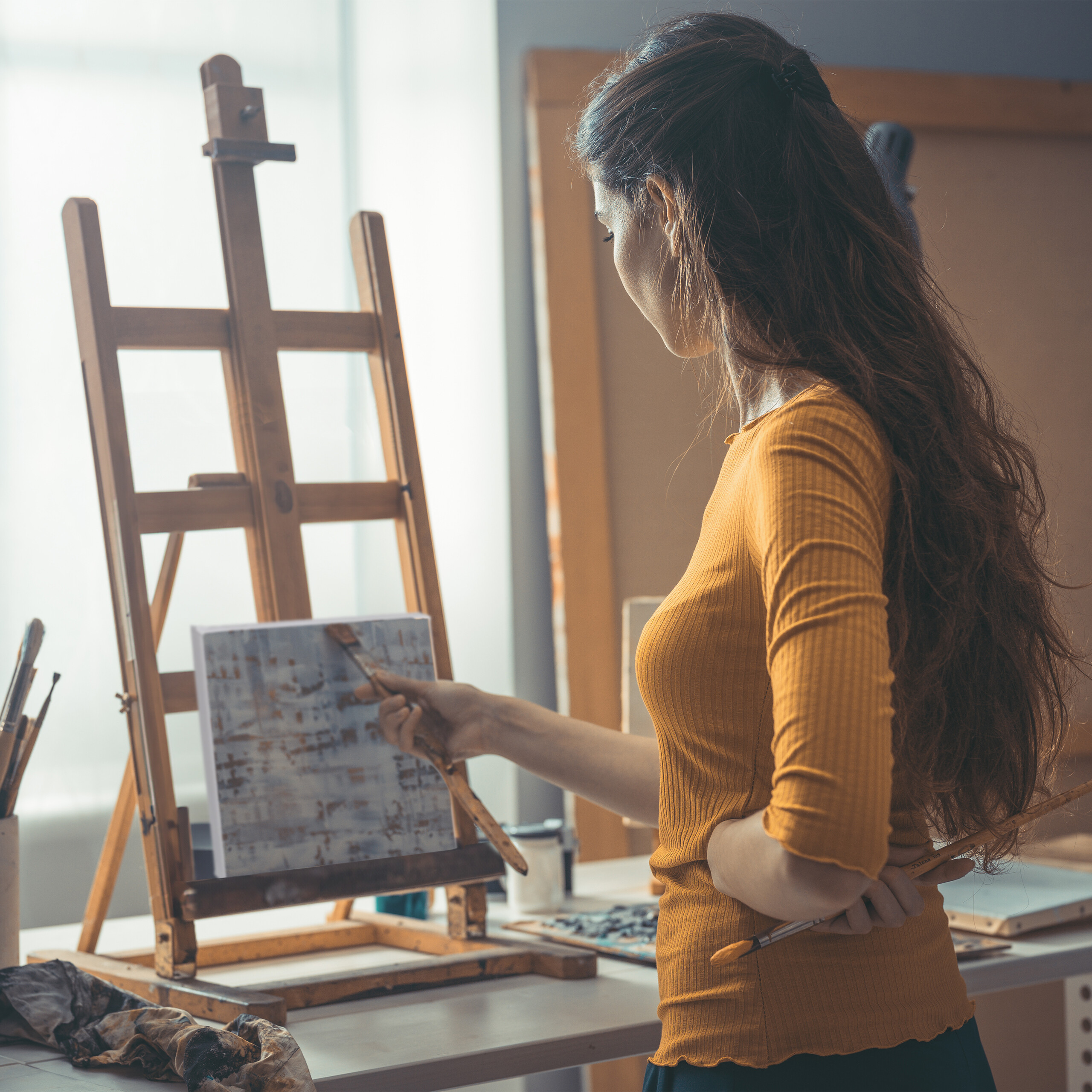Eine Künstlerin mit langem braunen Haar malt vor einer Holzeinheit auf einer Leinwand in einem Atelier.