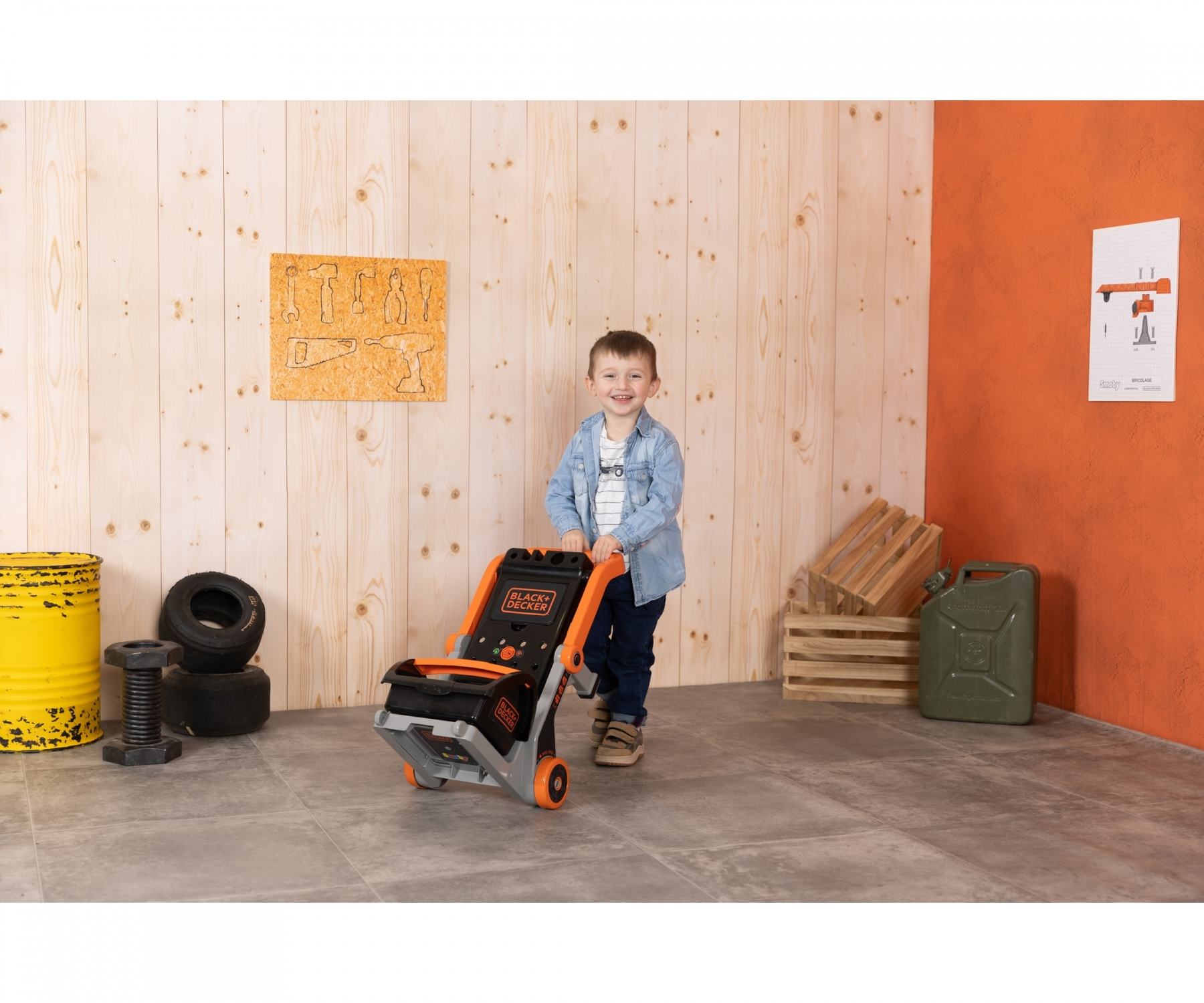 Ein kleiner Junge lächelt, während er in einer Werkstatt mit Holzwänden einen Spielzeug-Werkzeugwagen schiebt.