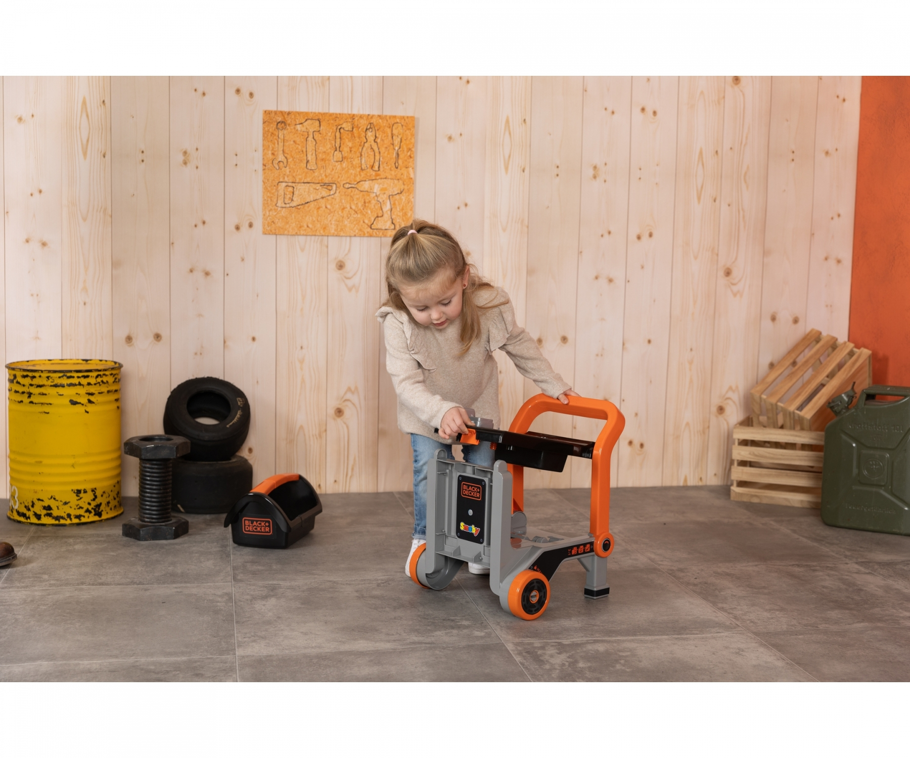 Ein junges Mädchen spielt mit einem orange-grauen Spielzeug-Werkzeugwagen in einer Werkstatt.