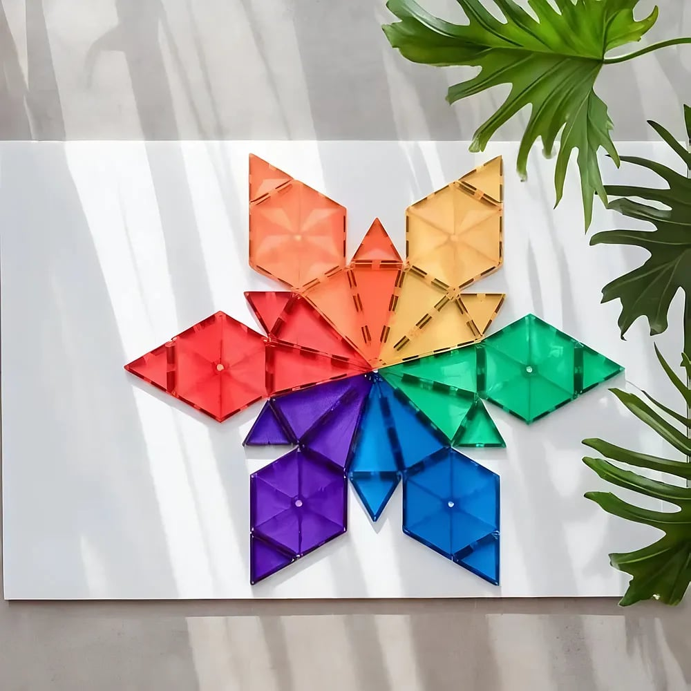 Ein buntes geometrisches Mandala aus durchsichtigen Magnetfliesen auf einer weißen Oberfläche, neben einem grünen Blatt.