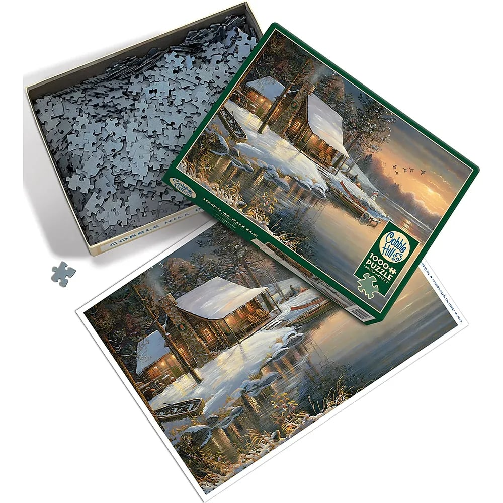 Ein offener Karton mit Puzzleteilen, der Deckel und ein fertiges Poster zeigen ein winterliches Blockhaus am See bei Sonnenuntergang.
