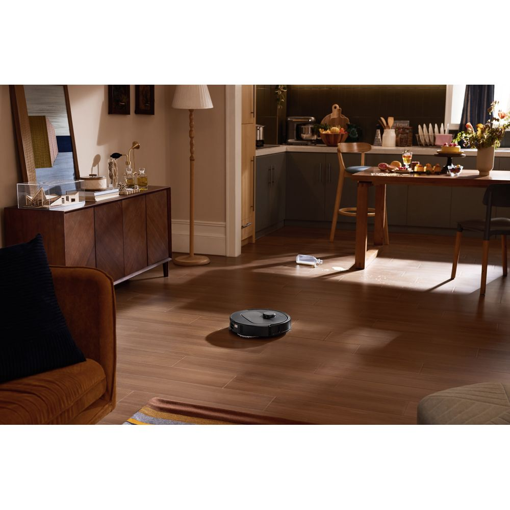 Un robot aspirapolvere nero pulisce il pavimento in legno del soggiorno, con una cucina visibile sullo sfondo.