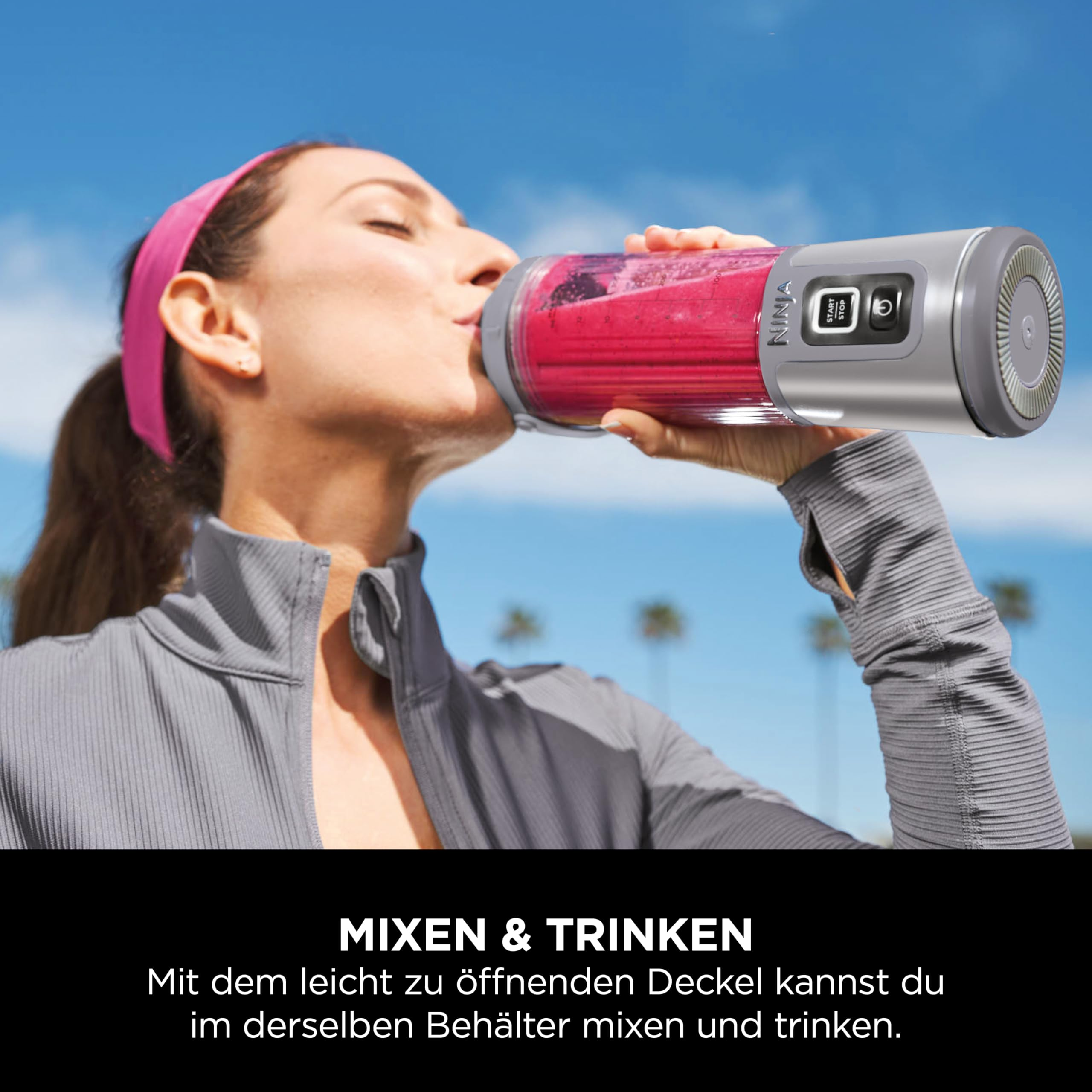 Eine Frau in Sportkleidung trinkt einen roten Smoothie aus einem Ninja-Mixbecher unter blauem Himmel.