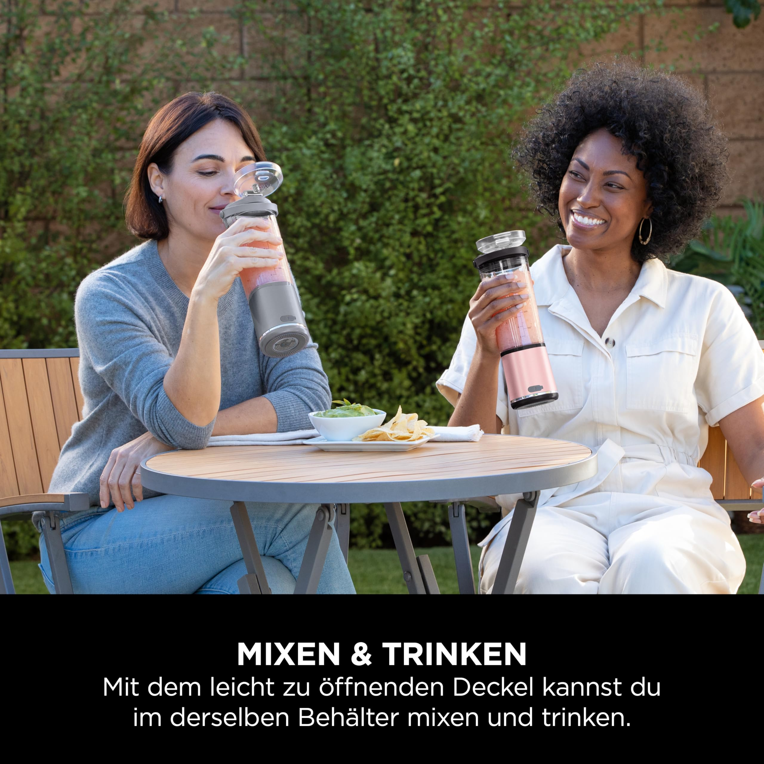 Zwei Frauen lächeln an einem Tisch im Freien und trinken aus tragbaren Mixern.