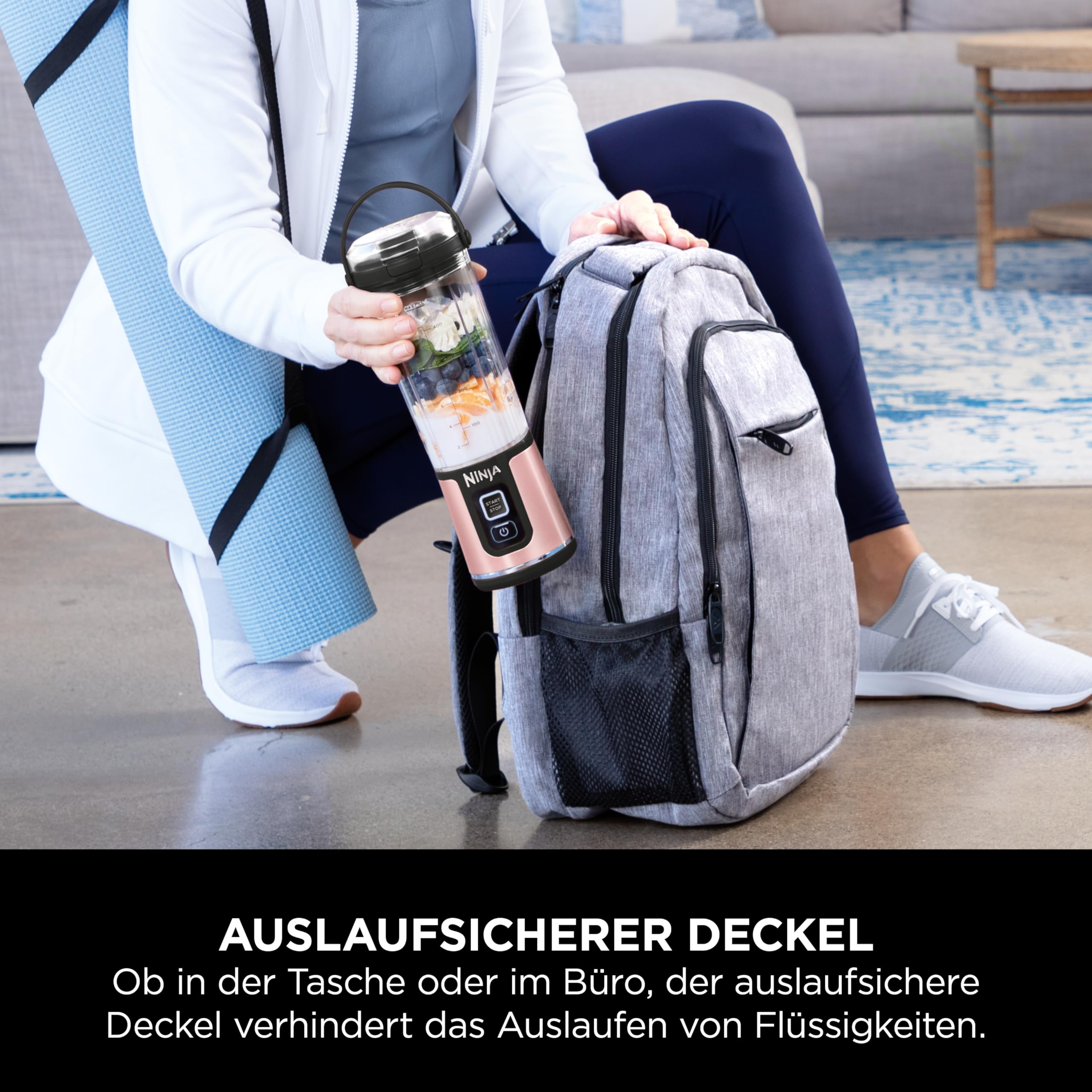 Eine Person hält einen tragbaren rosafarbenen Ninja-Mixer neben einem grauen Rucksack und einer blauen Yogamatte auf dem Boden.