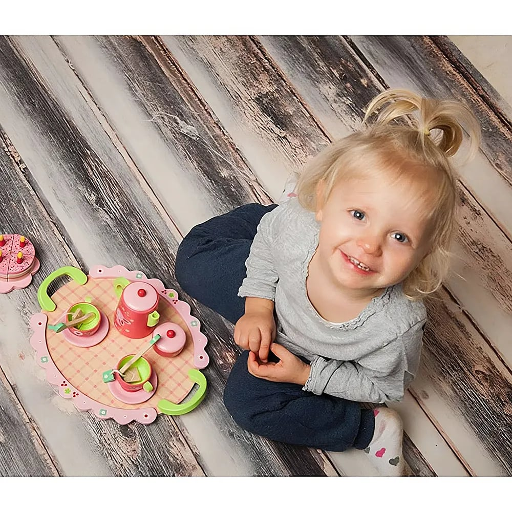 Petite fille aux tresses blondes assise sur un sol en bois, jouant avec une dinette rose.
