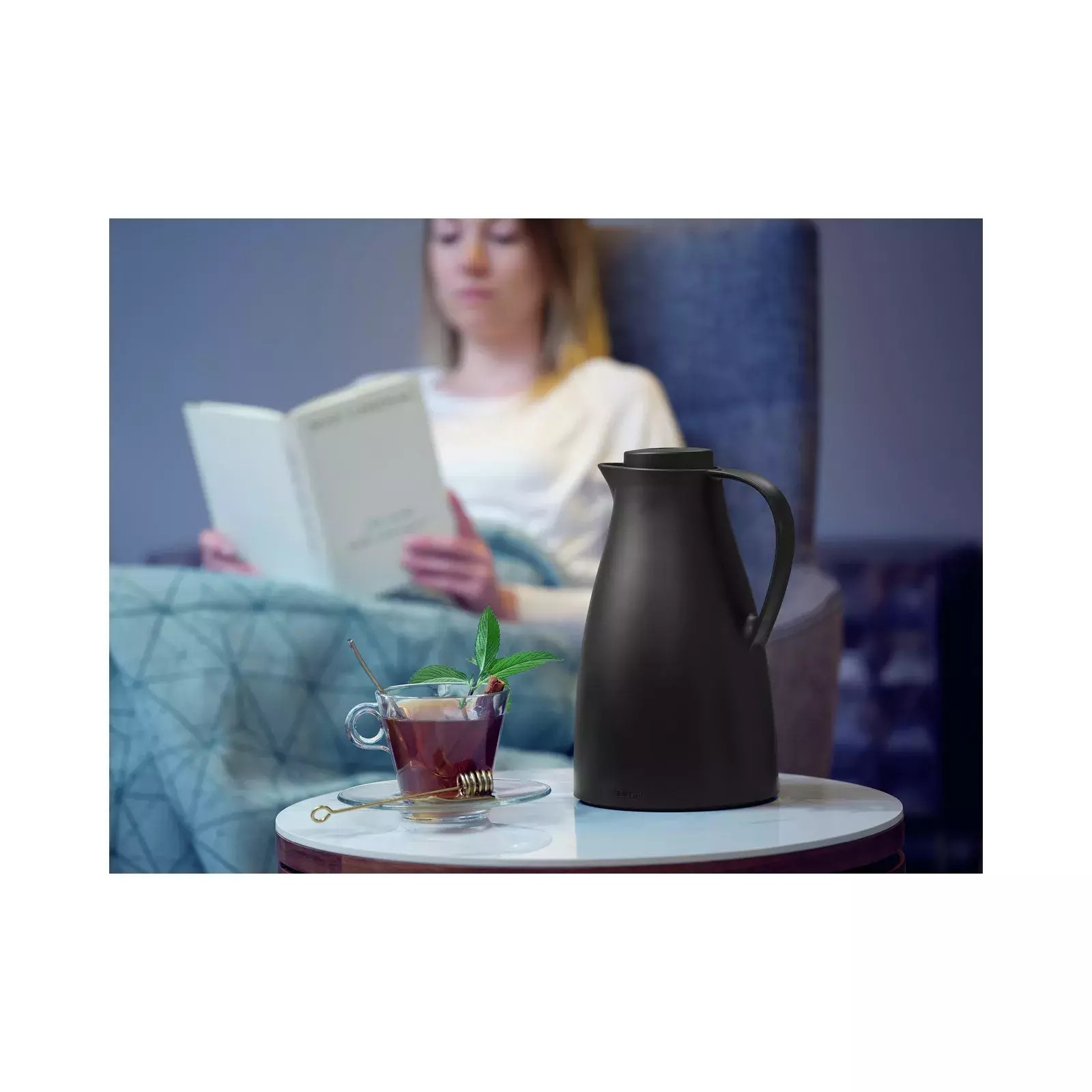 Eine schwarze Thermoskanne und eine Tasse Tee mit Minze stehen auf einem Tisch. Eine Frau liest im Hintergrund.