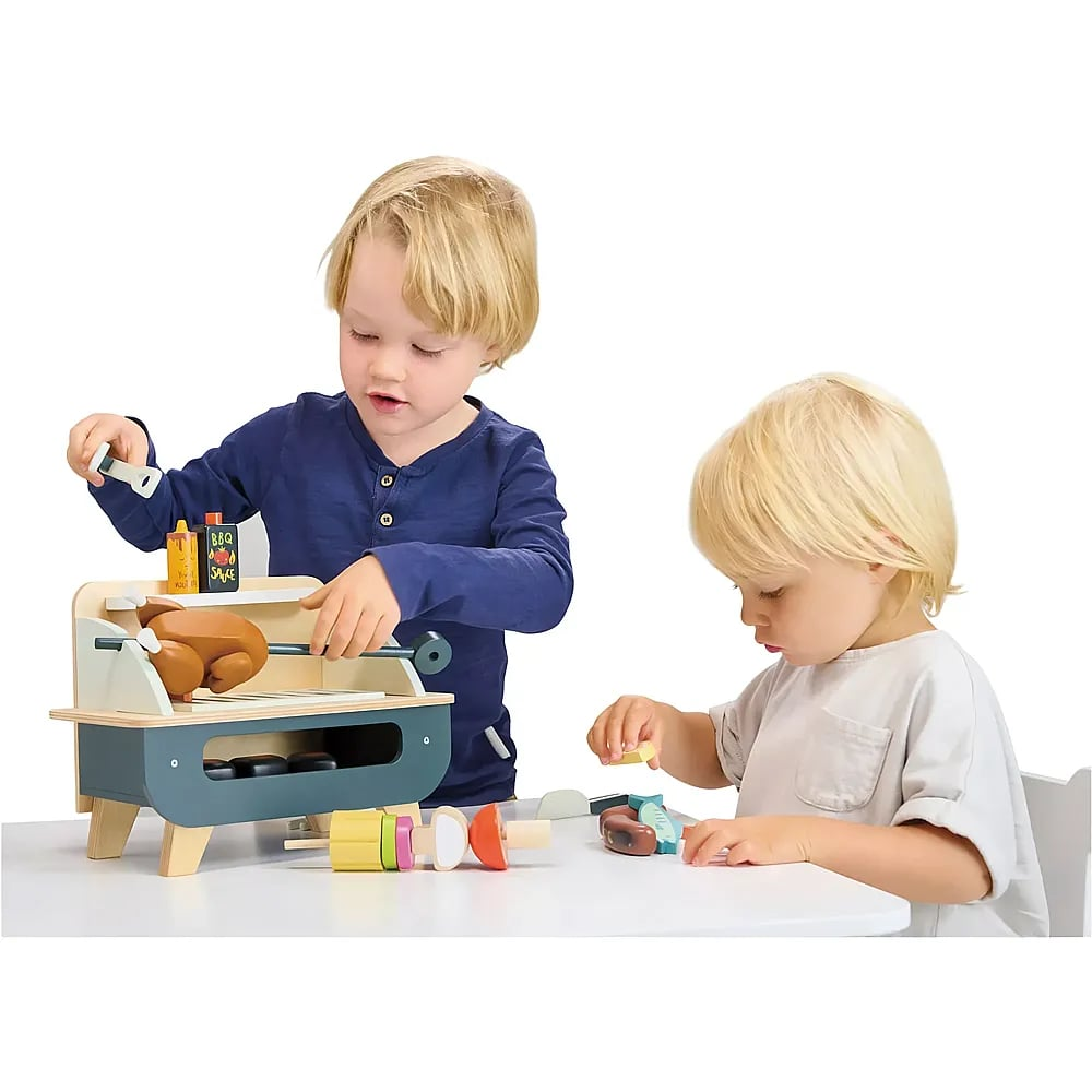 Zwei blonde Kinder spielen an einem weißen Tisch mit einem Holzkohlegrill und Spielzeug-Grillgut.