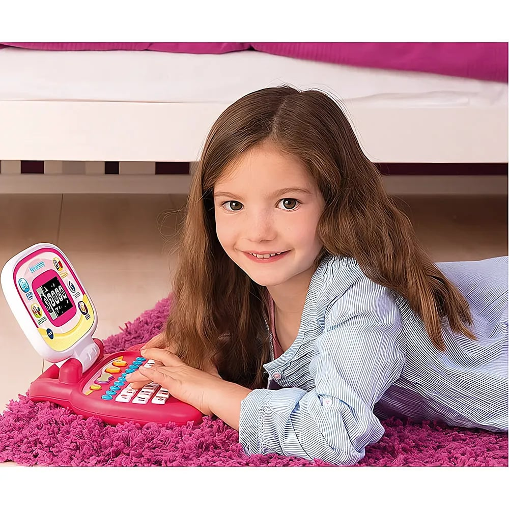 Ein junges Mädchen mit braunen Haaren liegt auf einem pinken Teppich und tippt auf einem kleinen rosa Spielzeuglaptop.