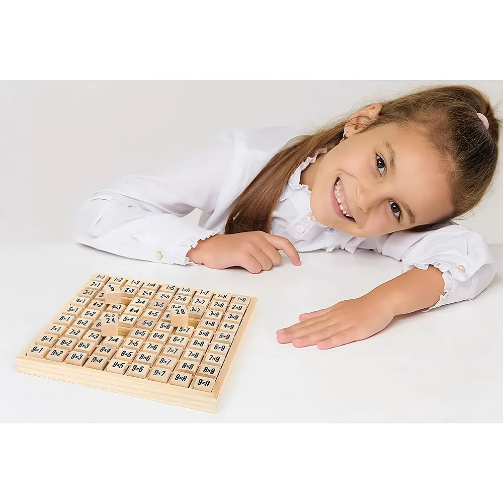 Ein lächelndes junges Mädchen in einem weißen Hemd posiert neben einem Einmaleins-Spiel auf einer weißen Oberfläche.