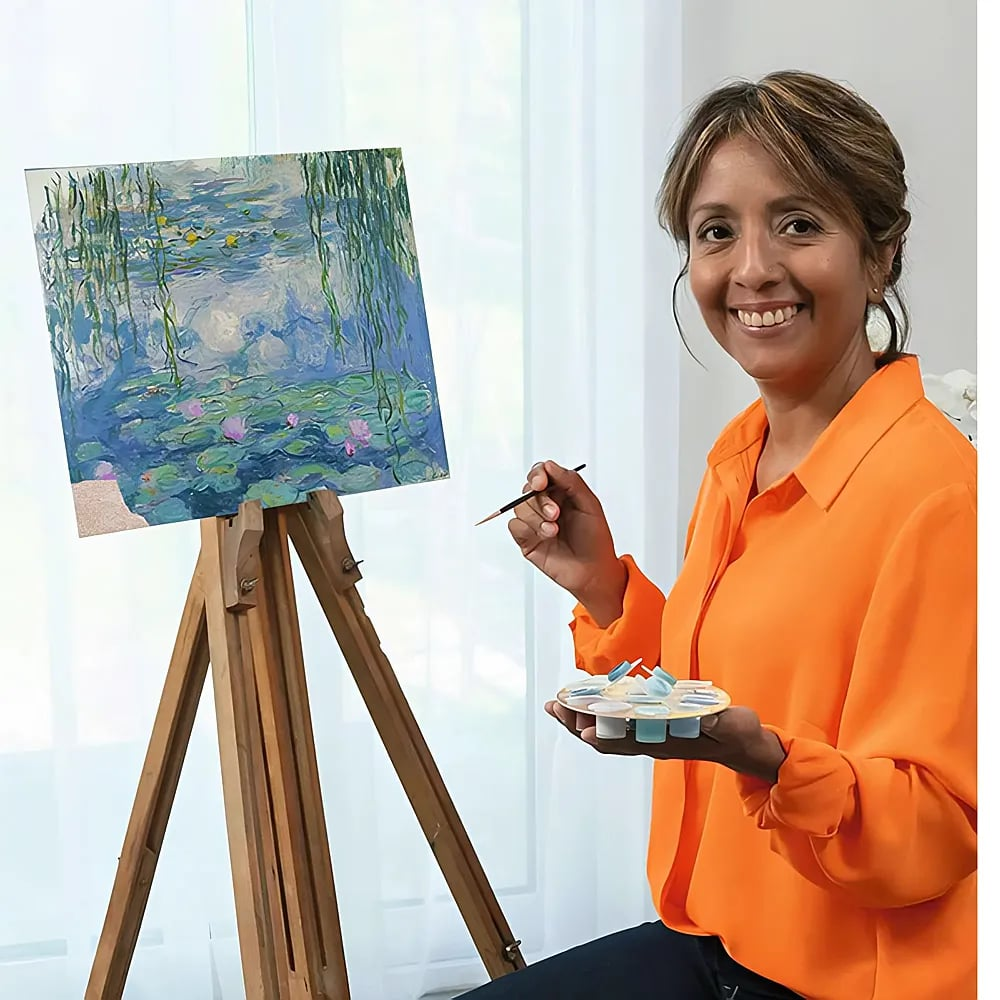 Una donna sorridente con una camicia arancione dipinge ninfee su un cavalletto.