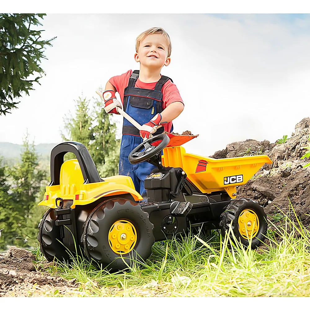 Ein kleiner Junge in Latzhose steht stolz draußen neben einem gelben JCB-Spielzeugmuldenkipper.