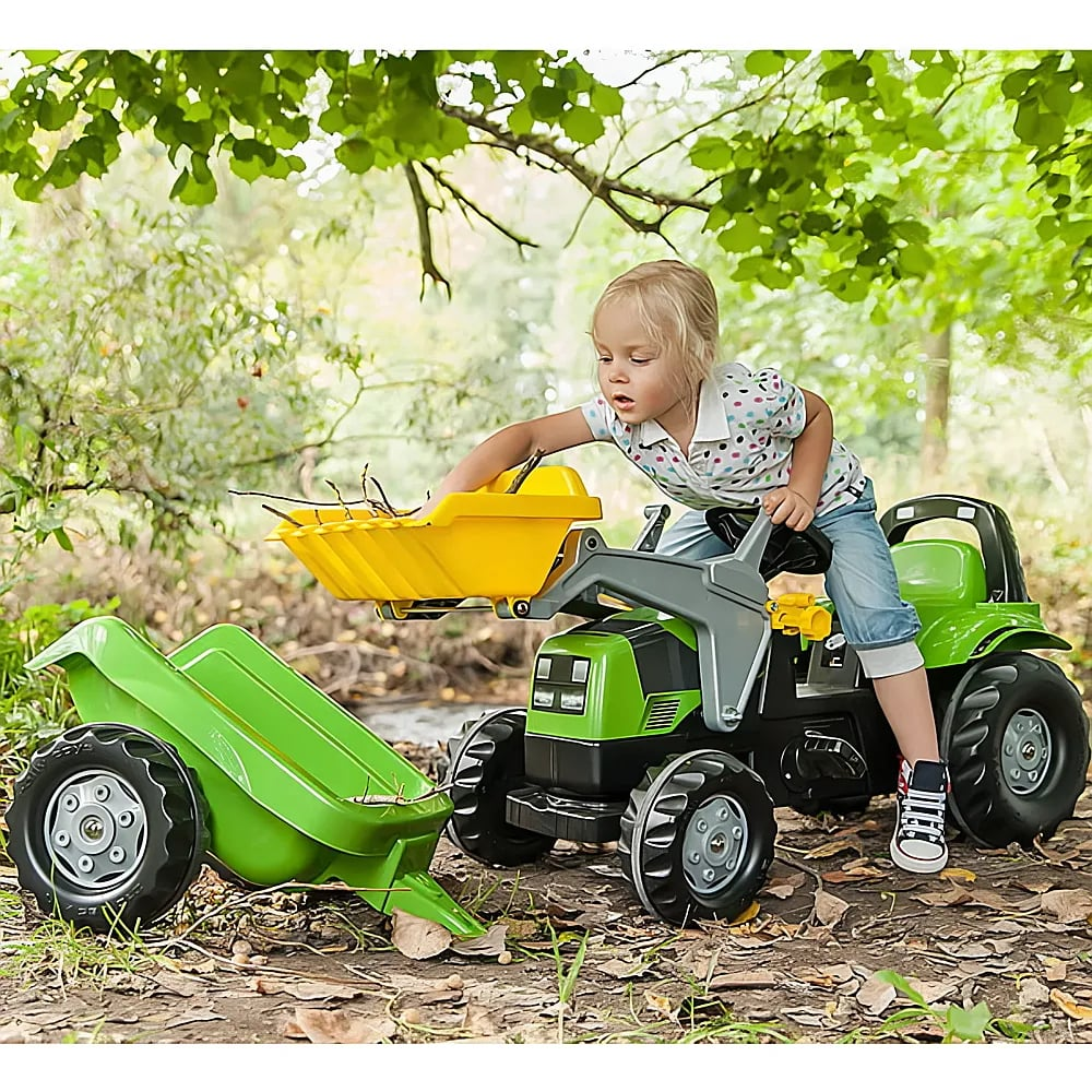 Ein Kleinkind in einem gemusterten Shirt sitzt auf einem grünen Spielzeugtraktor mit gelbem Lader im Wald.
