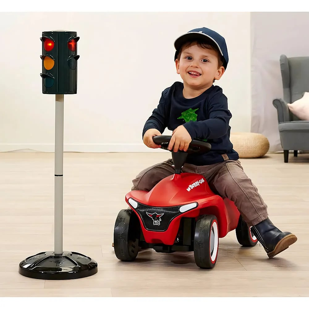 Ein lächelnder Junge sitzt auf einem roten Bobby-Car neben einer Spielzeug-Ampel.