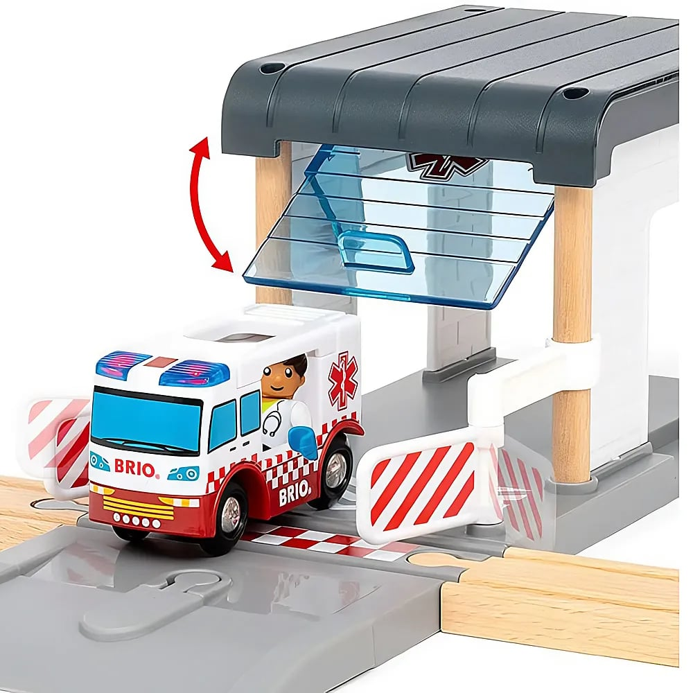 Ein Brio-Spielzeugkrankenwagen fährt aus einer geöffneten Garage mit roten und weißen Schranken auf Holzugschienen heraus.