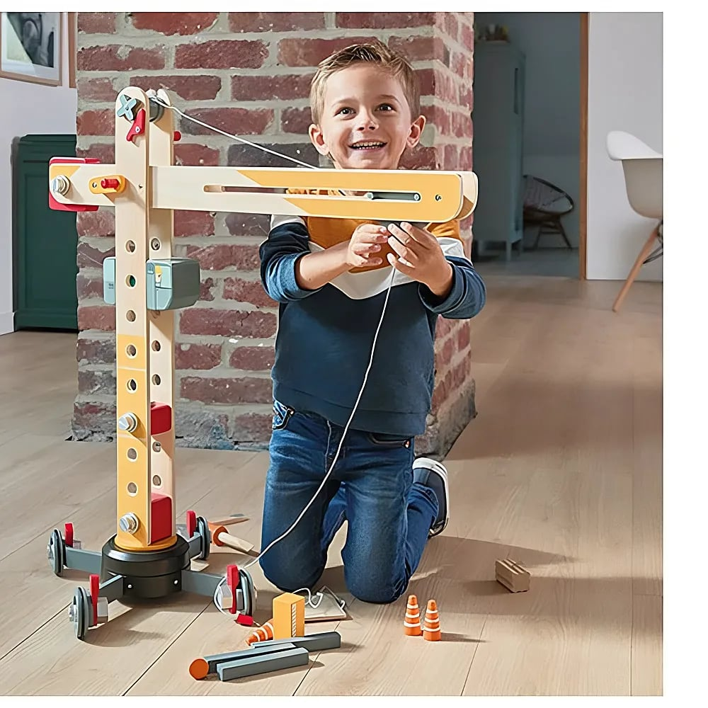 Ein lächelnder Junge kniet auf einem Holzboden und spielt mit einem großen Holzkran-Spielzeug.