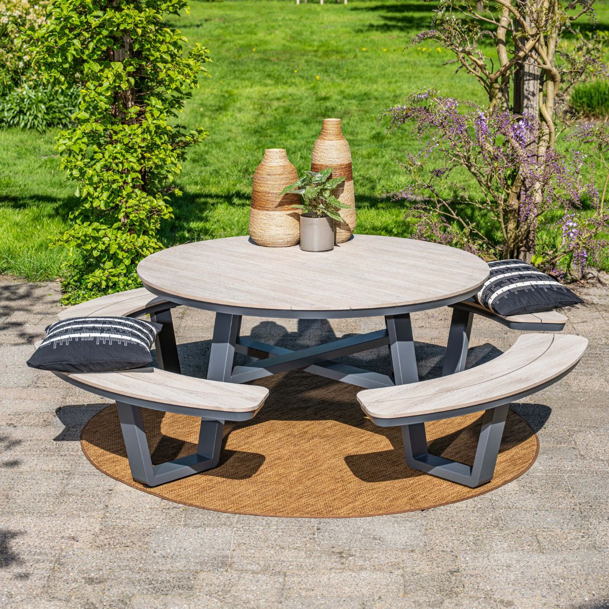Round patio dining set with wooden benches and cushions stands on a stone patio in a green garden.