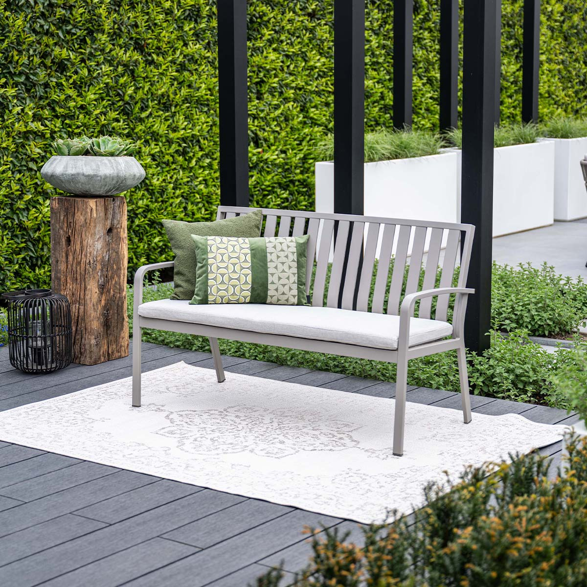A grey garden bench with pillows on a light rug on a patio with green plants in the background.